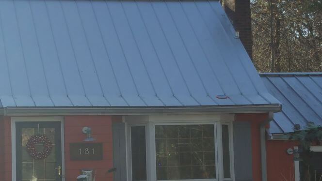 A pitched metal roof transitions with the metal bent and changing pitch. The metal is badly crimped and beginning to fail on the house in MA, CT, RI, or NH. 
