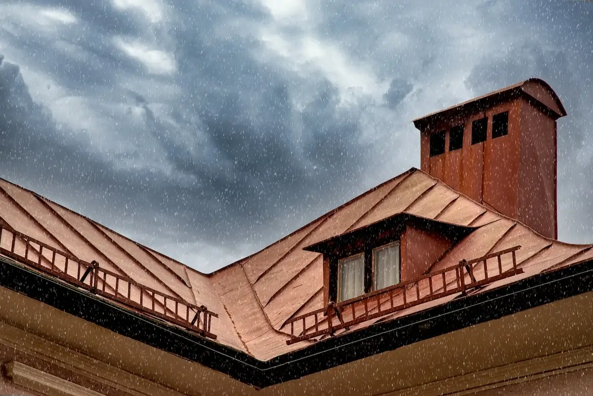 A copper-colored metal roof with a dormer window and chimney under a rain shower.